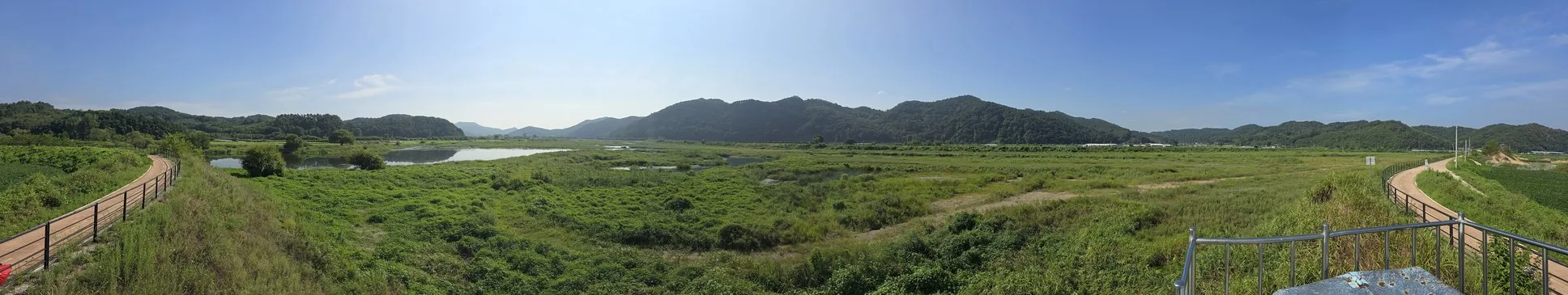 A real cycling and walking trail through a green wetland valley, boardwalk railing in the foreground, wooded hills in the distance.
